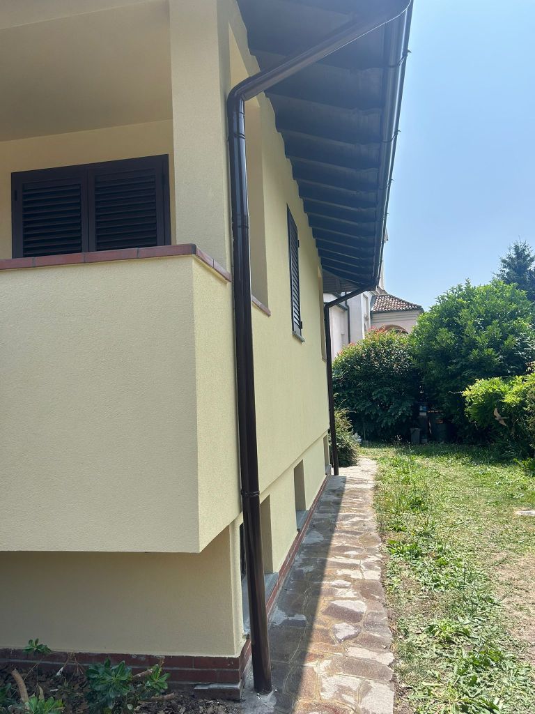 Lato di un edificio con parete gialla, finestra con persiane scure e un tubo di gronda. Presente un sentiero in pietra e vegetazione circostante.