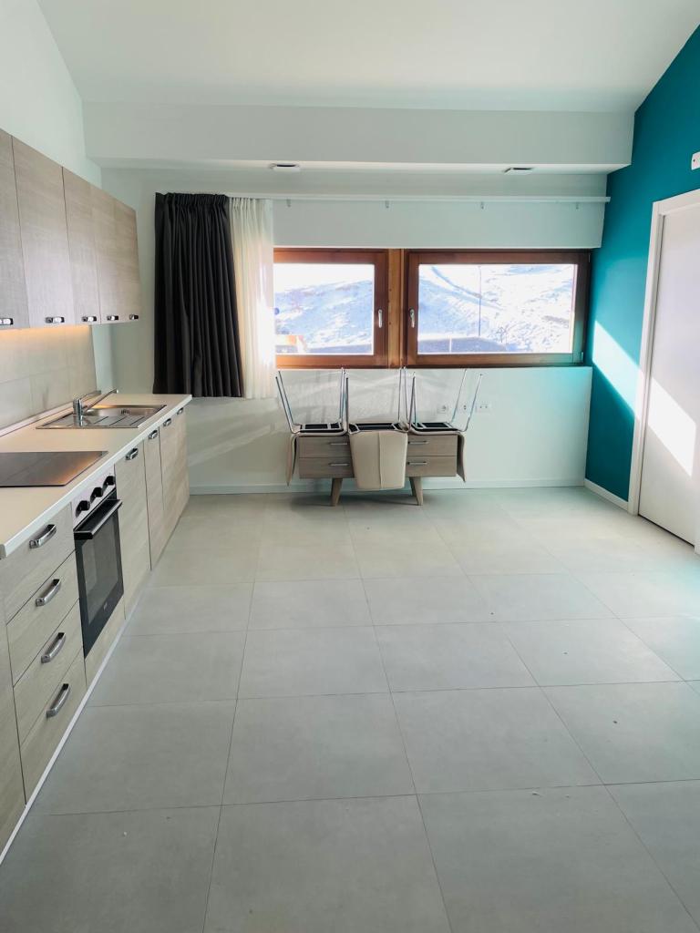 Cucina moderna con armadi in legno chiaro, piano cottura e forno. Due finestre con tende bianche che offrono una vista esterna. Area pranzo con sedie impilate.