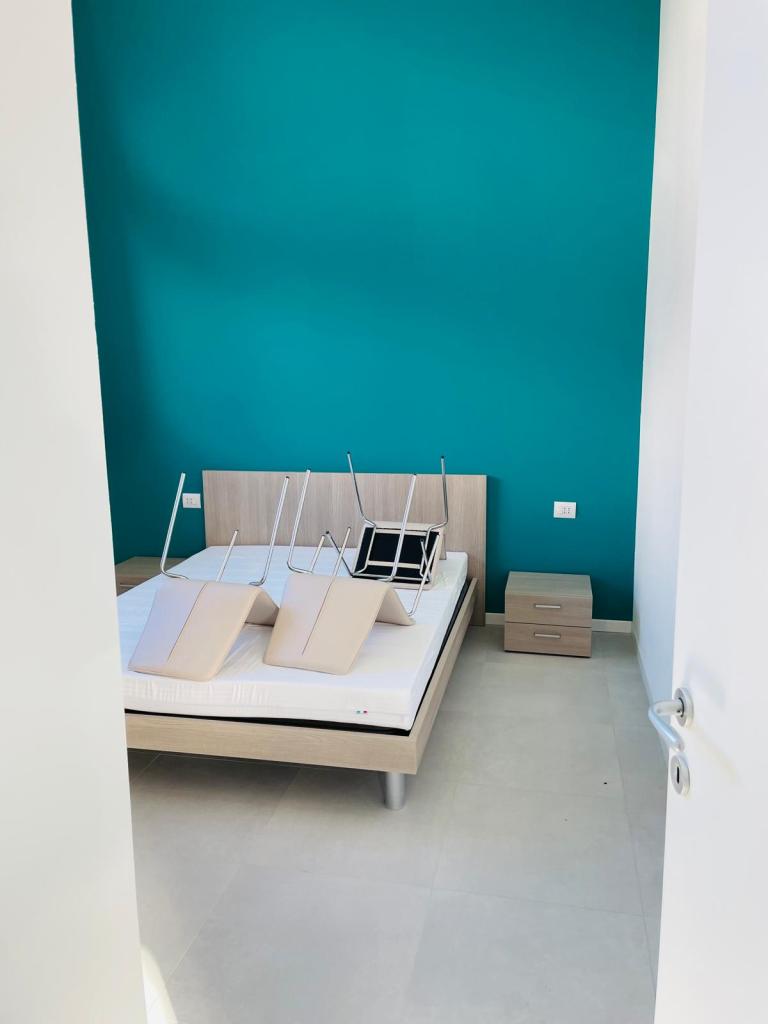 Una camera da letto moderna con parete verde e un letto con materasso bianco. Alcuni oggetti sono appesi sopra il letto e c'è un comodino accanto.