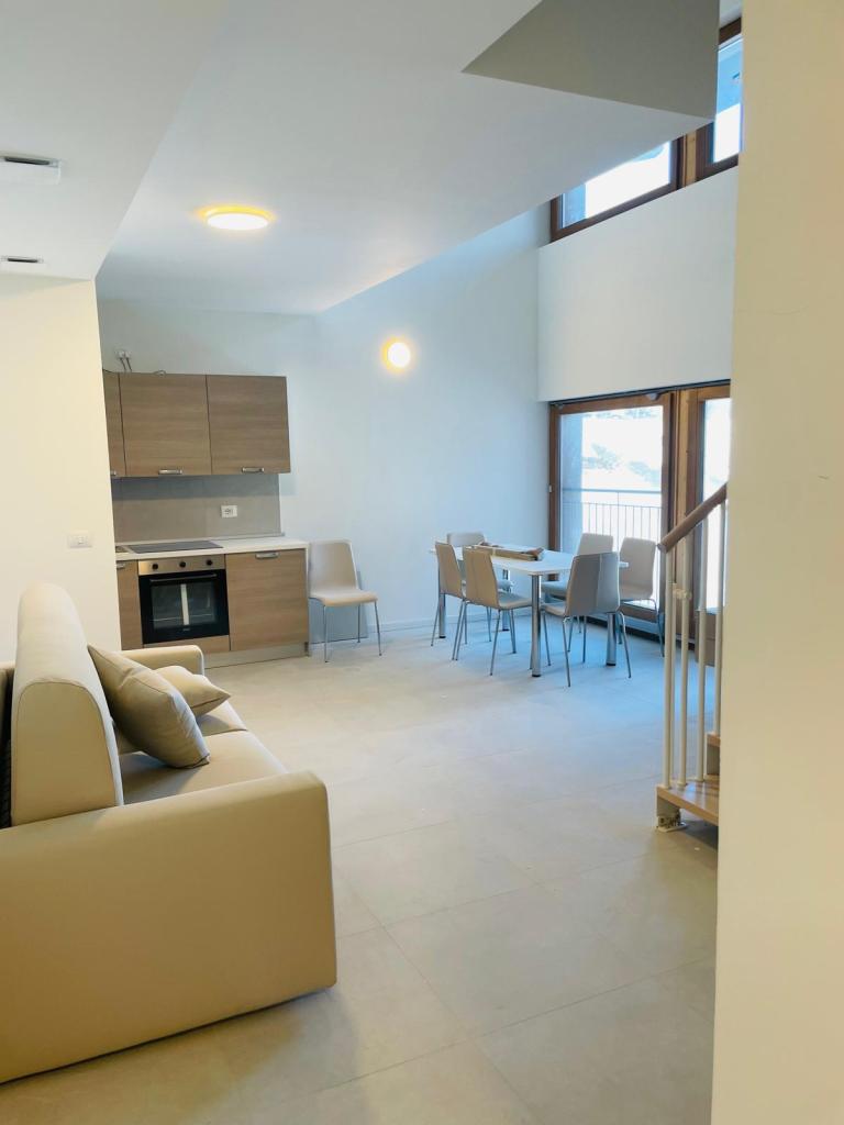 Interno di un soggiorno moderno con cucina e zona pranzo. Divano chiaro a sinistra, tavolo e sedie a destra, finestre luminose.